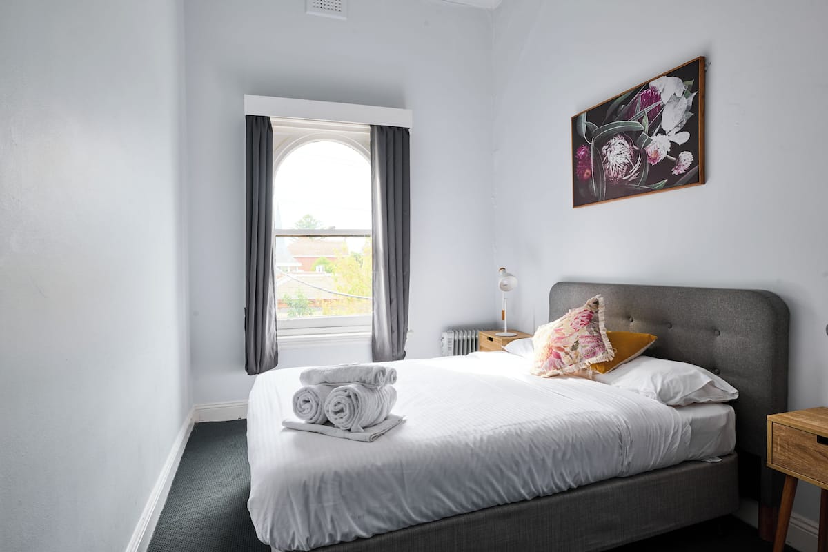 Una cama bien hecha con toallas enrolladas, un cabecero gris, una mesita de noche con lámpara y una ventana con cortinas.