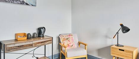 A room with a wooden desk, a chair with a floral cushion, a bedside table with a lamp, and a painting on the wall.