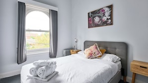 A neatly made bed with rolled towels, a bedside table with a lamp, and a window with curtains. - College Lawn Hotel - Hostel (Prahran)