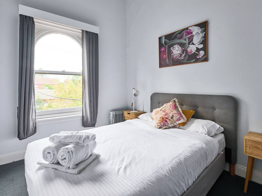 A neatly made bed with rolled towels, a bedside table with a lamp, and a window with curtains.