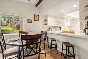 A modern kitchen with white cabinets, a dining area with wooden chairs and a table, and a view of a green landscape through large windows. - Cozy, Comfortable on Oak Creek Golf Course! (Sedona)