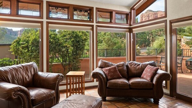 A spacious room with large windows, a brown leather sofa, and a matching ottoman. - Home Surrounded by Lush Landscape + Mtn Views! (Sedona)