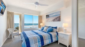 A bedroom with a large bed, two bedside tables with lamps, a chair, a ceiling fan, and a view of the ocean.
