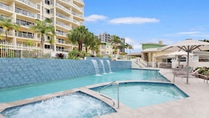 A hotel pool area with a hot tub, a waterfall feature, and a lounge area with umbrellas.