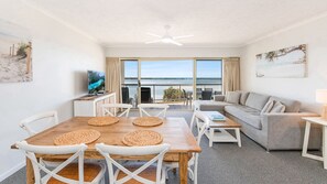 A modern living room with a dining area, a sofa, a television, and a view of the sea.