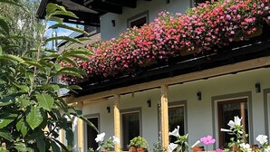 Un immeuble de deux étages doté de balcons ornés de fleurs roses, entouré d’une végétation luxuriante et de fleurs blanches.