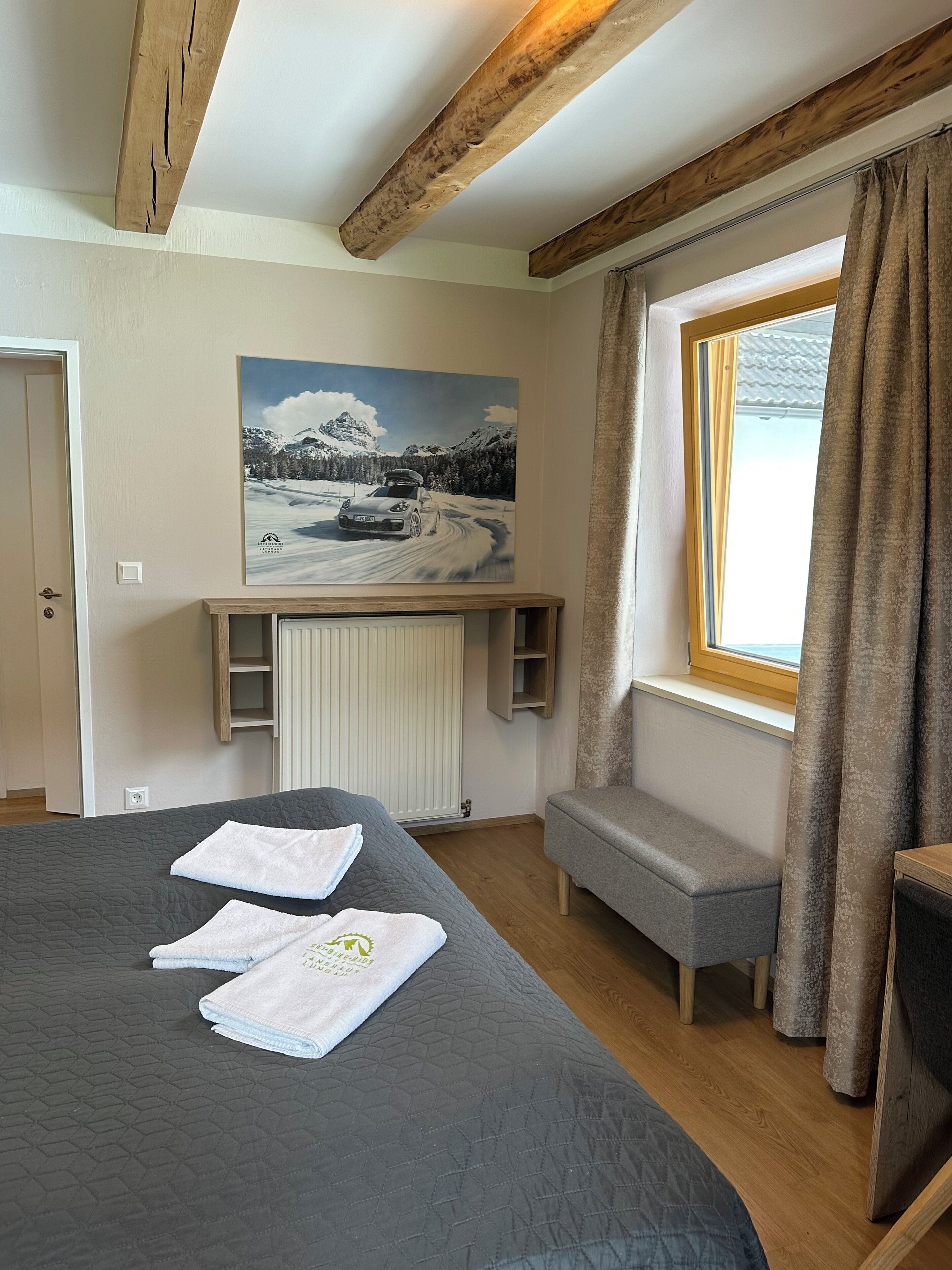 A bedroom with a large bed, a window with curtains, a painting on the wall, and a built-in bench.