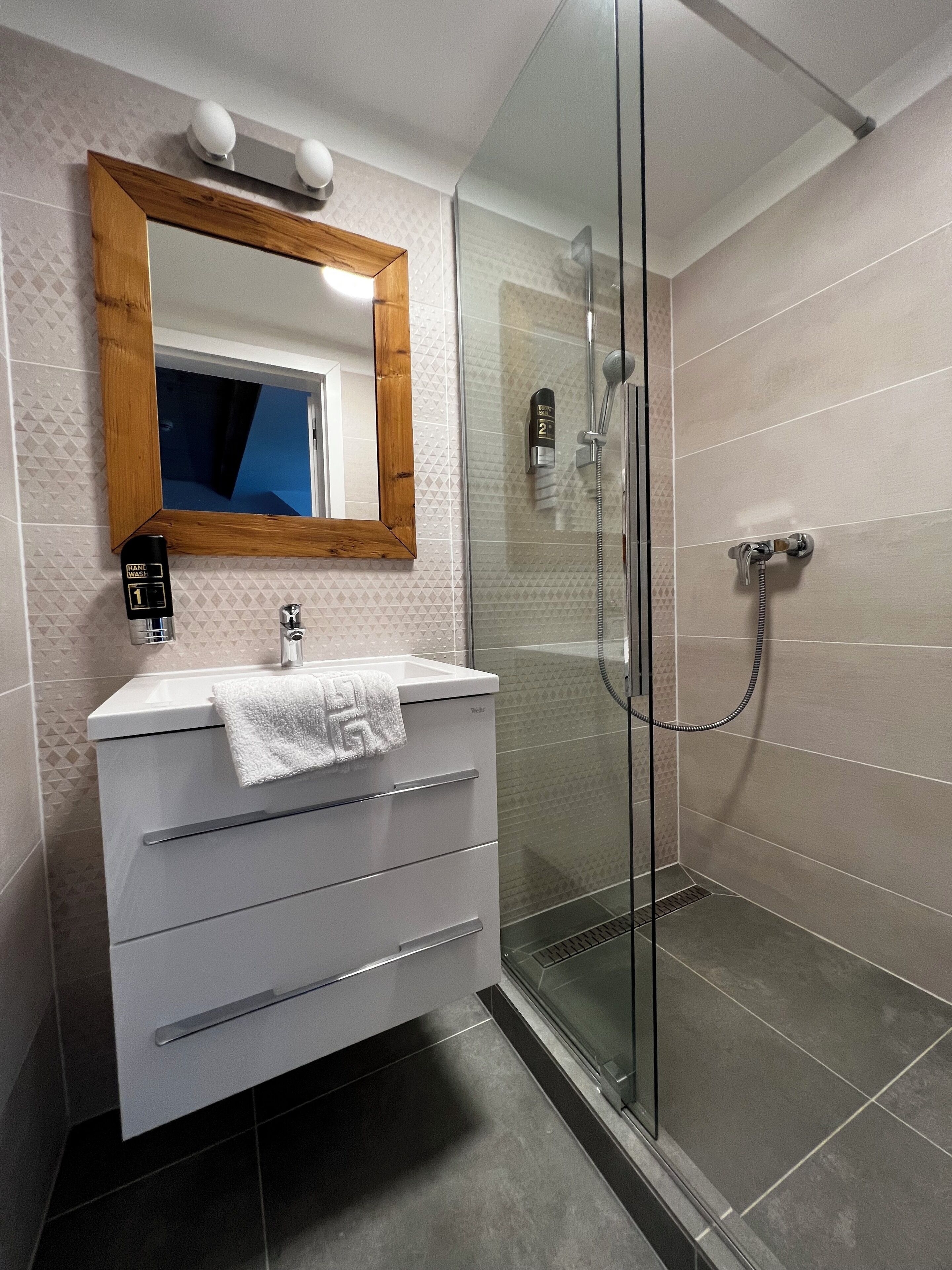 A modern bathroom with a glass shower enclosure, a wooden-framed mirror, and a white vanity with a towel.