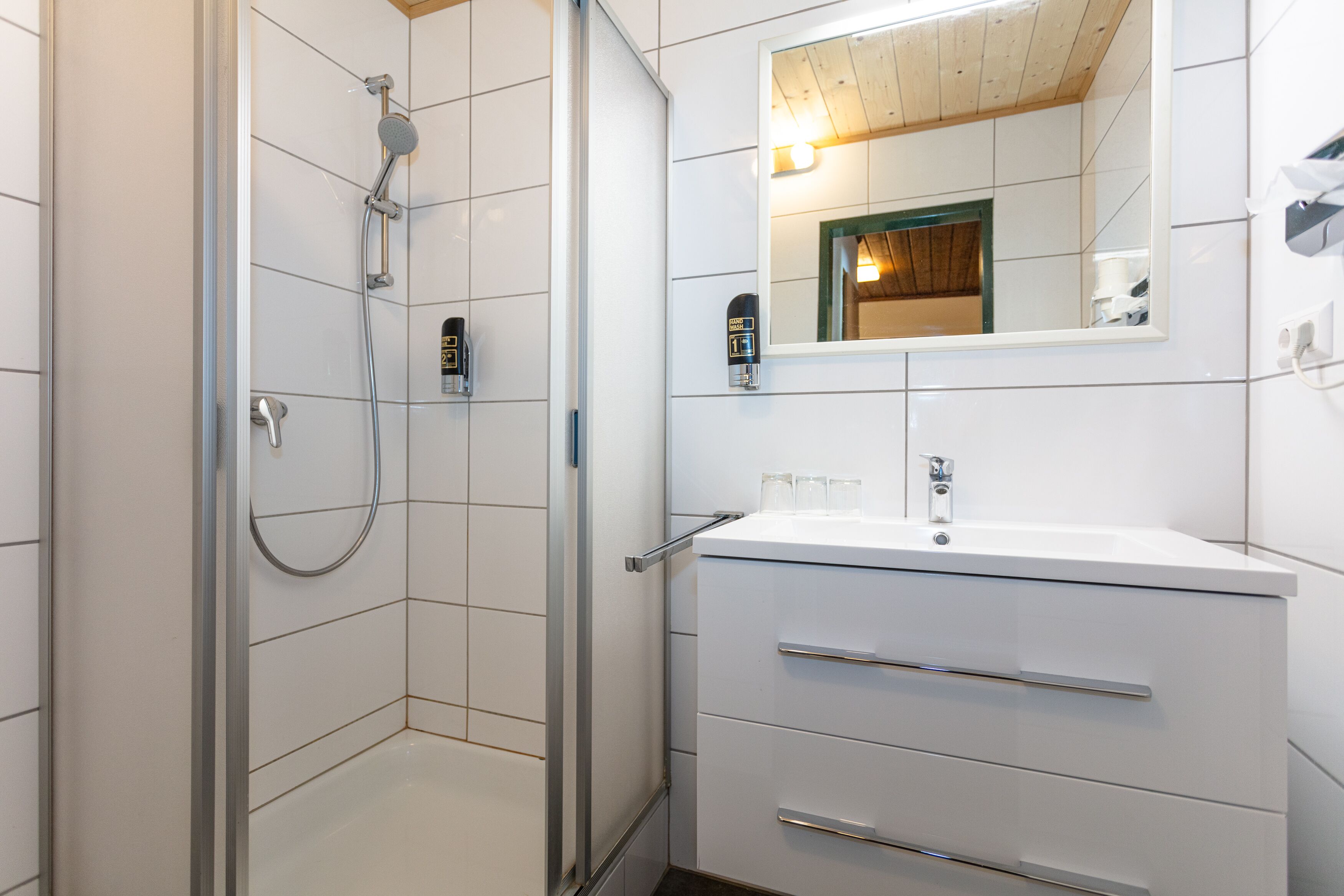 Un baño moderno con cabina de ducha de vidrio, un lavamanos blanco con un espejo encima y techo de madera.