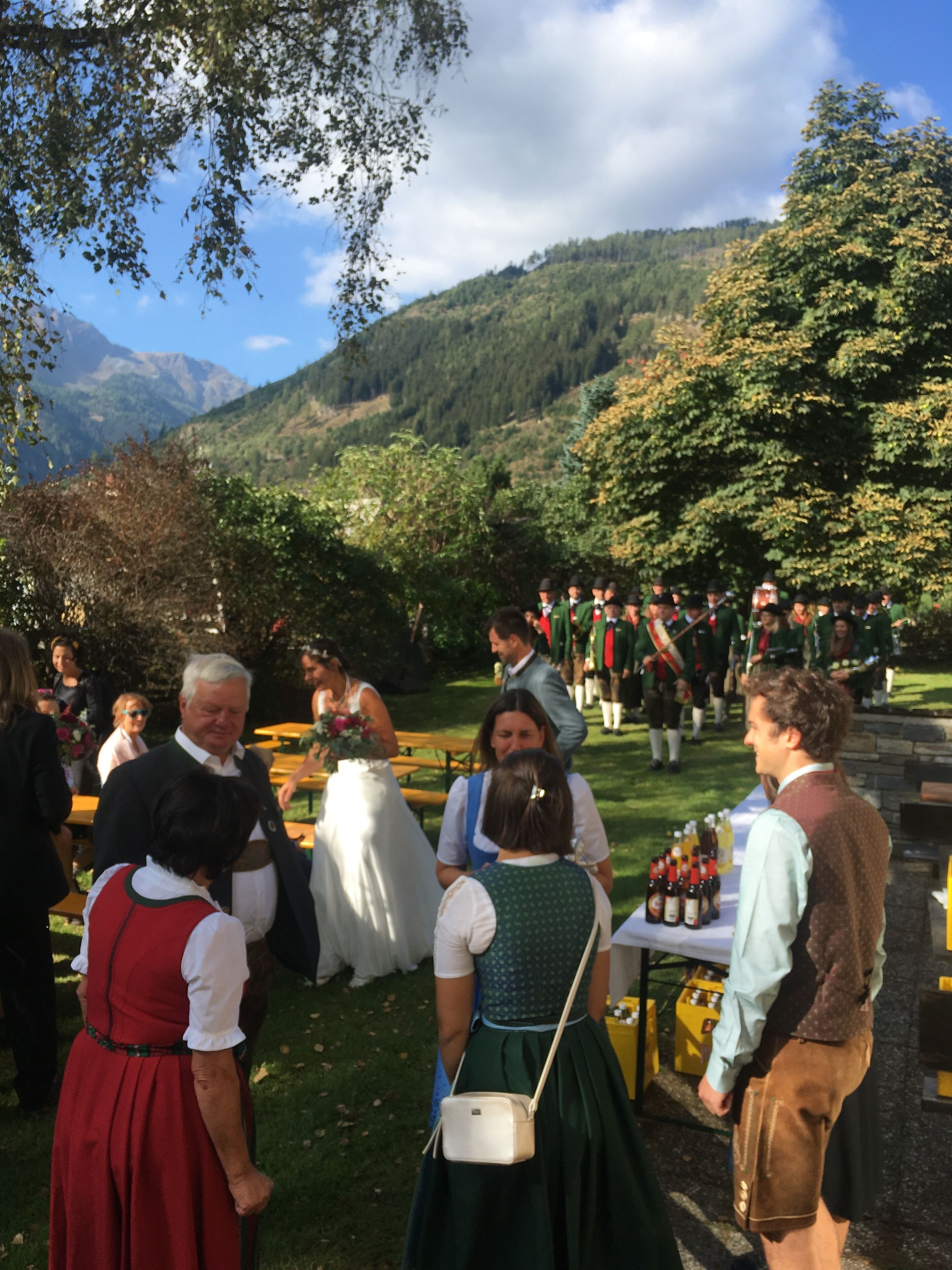 A group of people in traditional attire gathered outdoors, with a mountainous backdrop and a table with bottles.