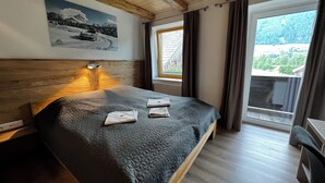Une chambre à coucher avec une tête de lit en bois, un lit recouvert d’une courtepointe, une table de chevet, une lampe fixée au mur, une fenêtre avec des rideaux et une vue sur un paysage montagneux.
