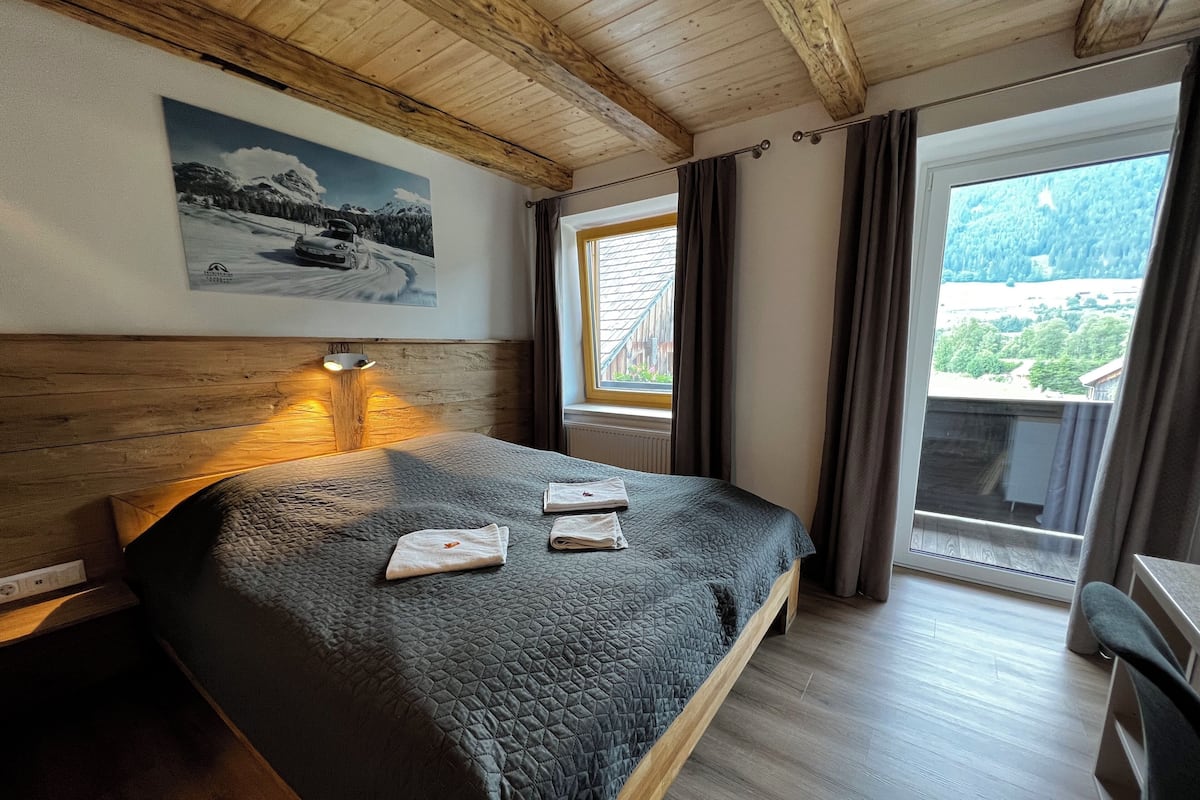 A bedroom with a wooden headboard, a bed with a quilted cover, a nightstand, a wall-mounted lamp, a window with curtains, and a view of a mountainous landscape.
