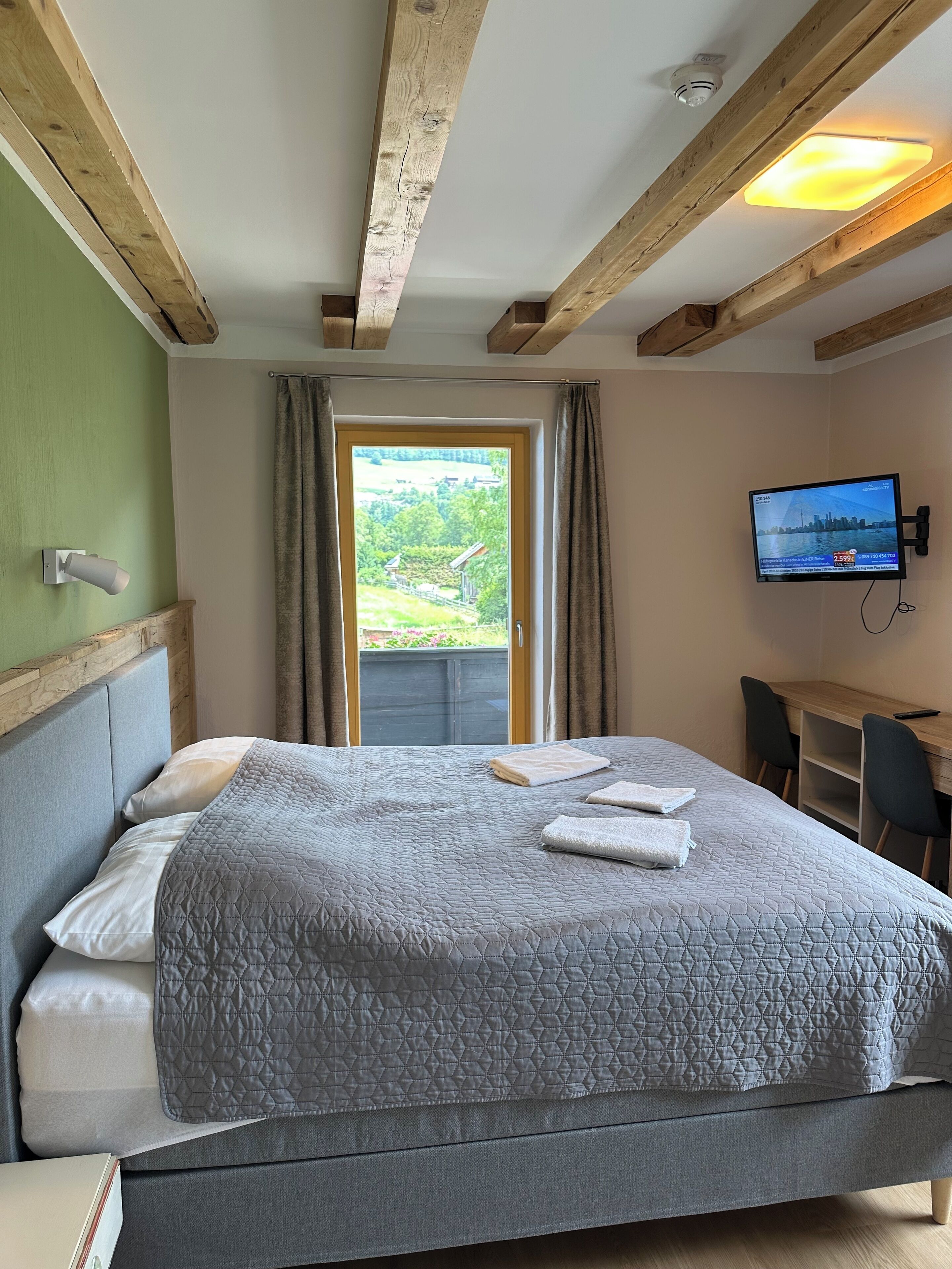 Una habitación de hotel con cama, escritorio, televisión y ventana con cortinas.