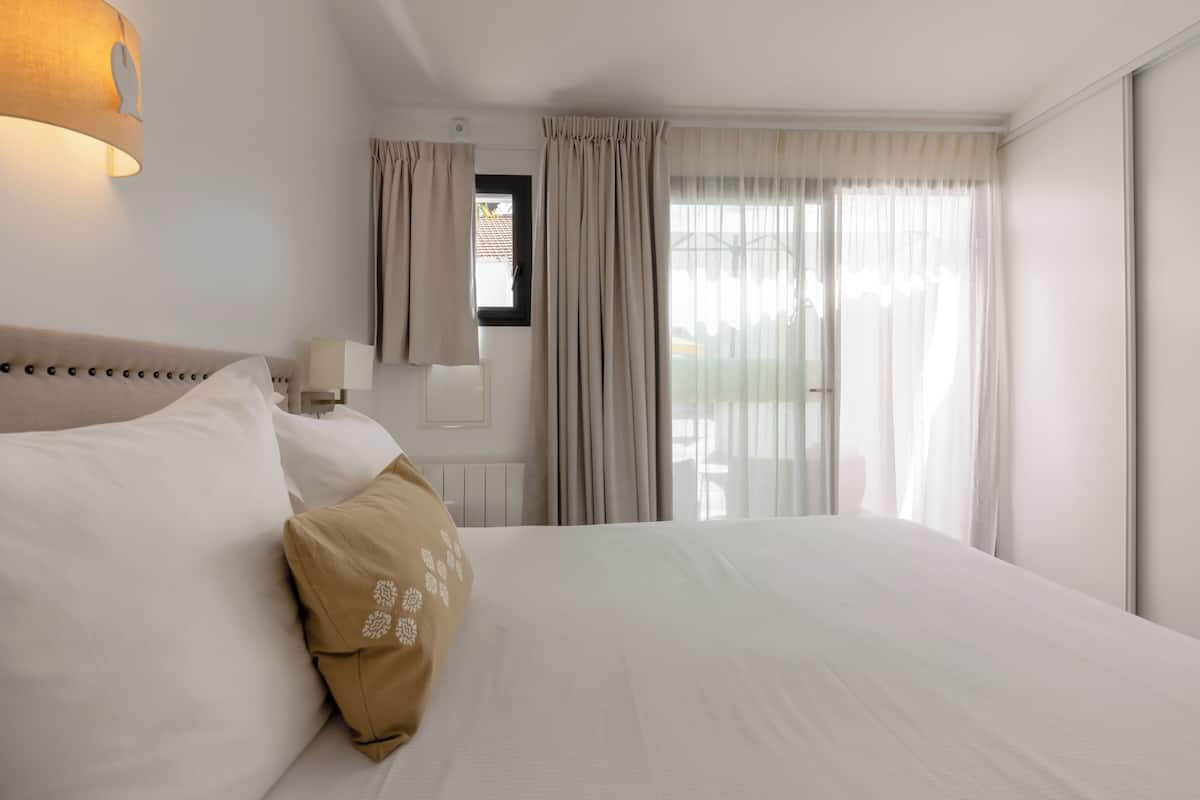 A hotel room with a large bed, a bedside table with a lamp, a window with curtains, and a view of the outside.