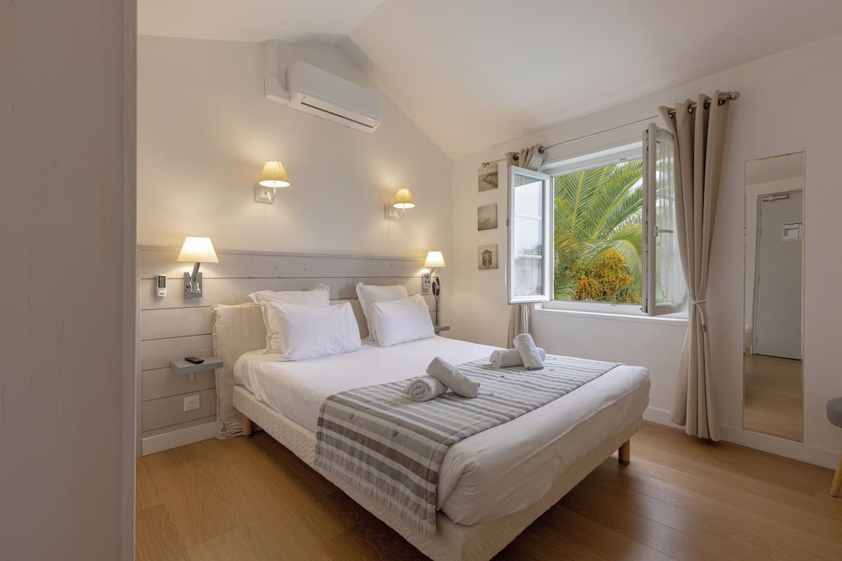 A bedroom with a bed, bedside lamps, a window with curtains, and a wall-mounted air conditioner.