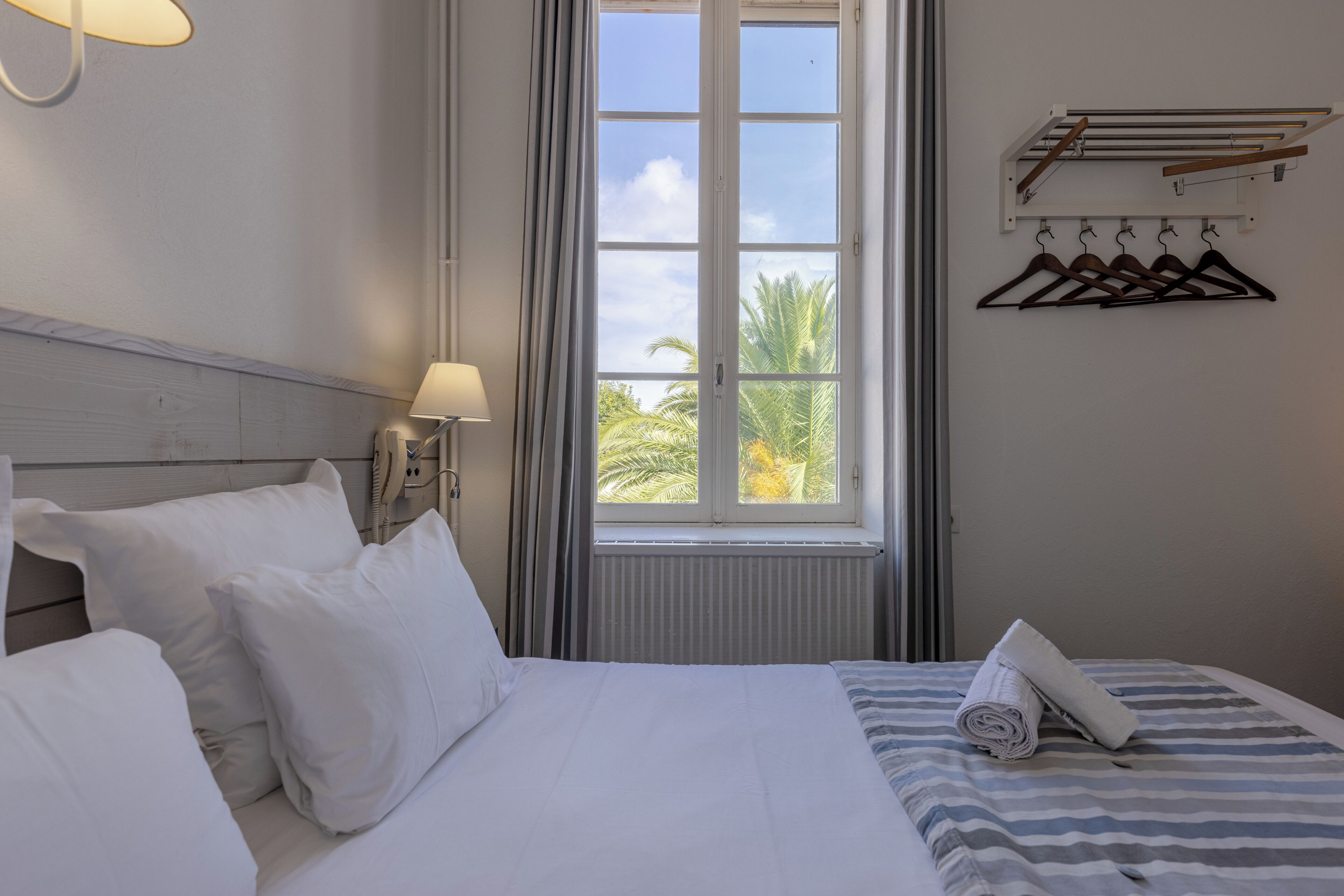 A hotel room with a bed, pillows, a window with a view of palm trees, a towel, and a coat rack with hangers.