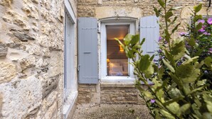 A stone building with a window featuring blue shutters, a small step leading to the entrance, and a plant with purple flowers in the foreground. - Hotel Le Square (Saint-Pierre-d'Oleron)