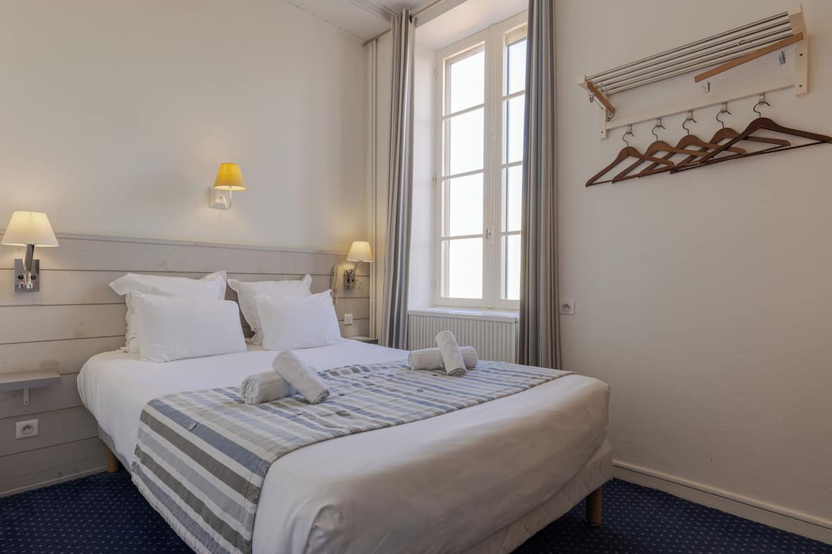 A neatly made bed with white linens, a striped blanket, and rolled towels. There are two bedside lamps, a window with curtains, and a wall-mounted drying rack with hangers.