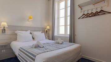Ein ordentlich bezogenes Bett mit weißen Leinen, einer gestreiften Decke und zusammengerollten Handtüchern. Zwei Nachttischlampen, ein Fenster mit Vorhängen und ein an der Wand befestigter Wäscheständer mit Kleiderbügeln sind ebenfalls zu sehen.