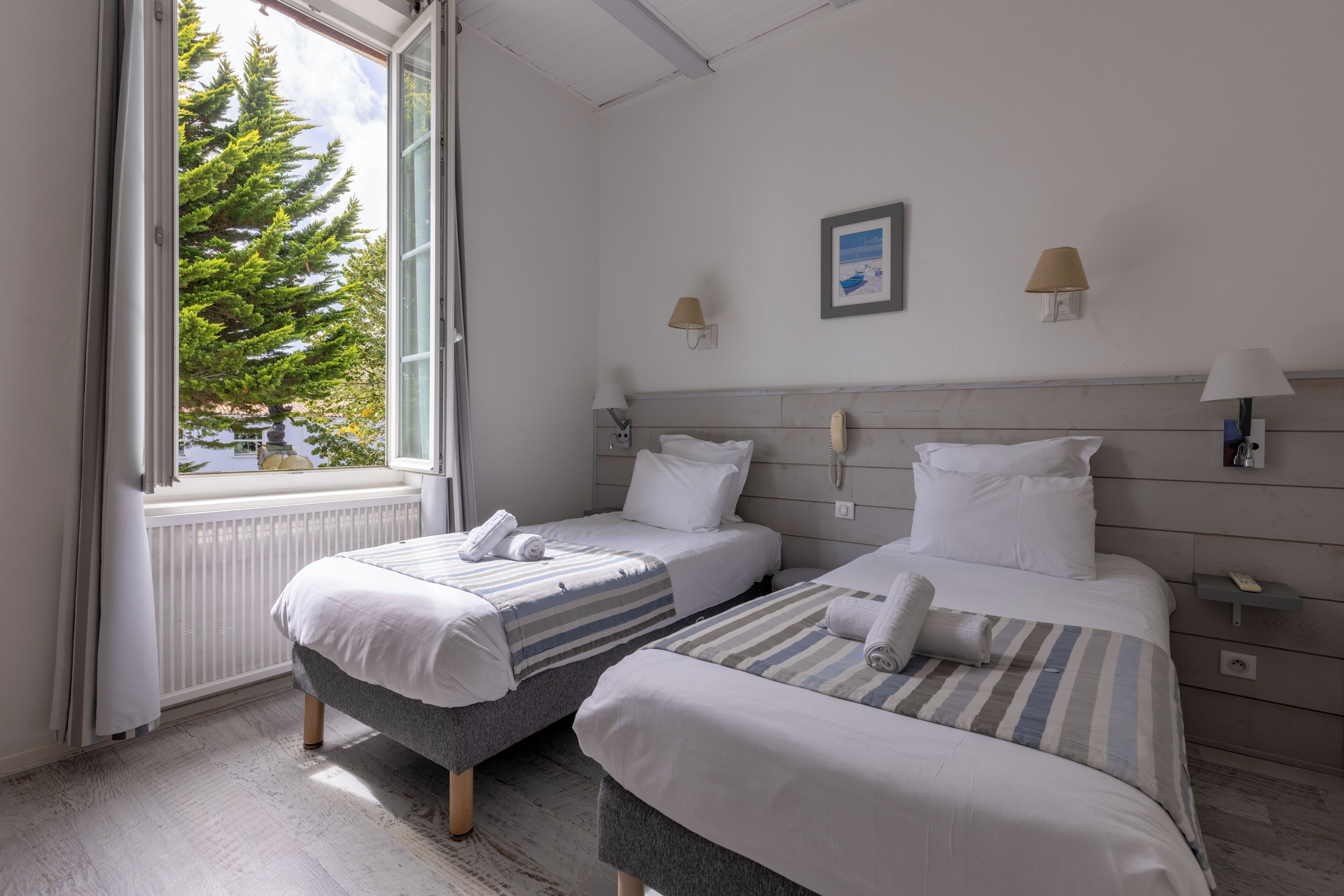 A hotel room with two beds, a window with a view of a tree, and a wall-mounted lamp.