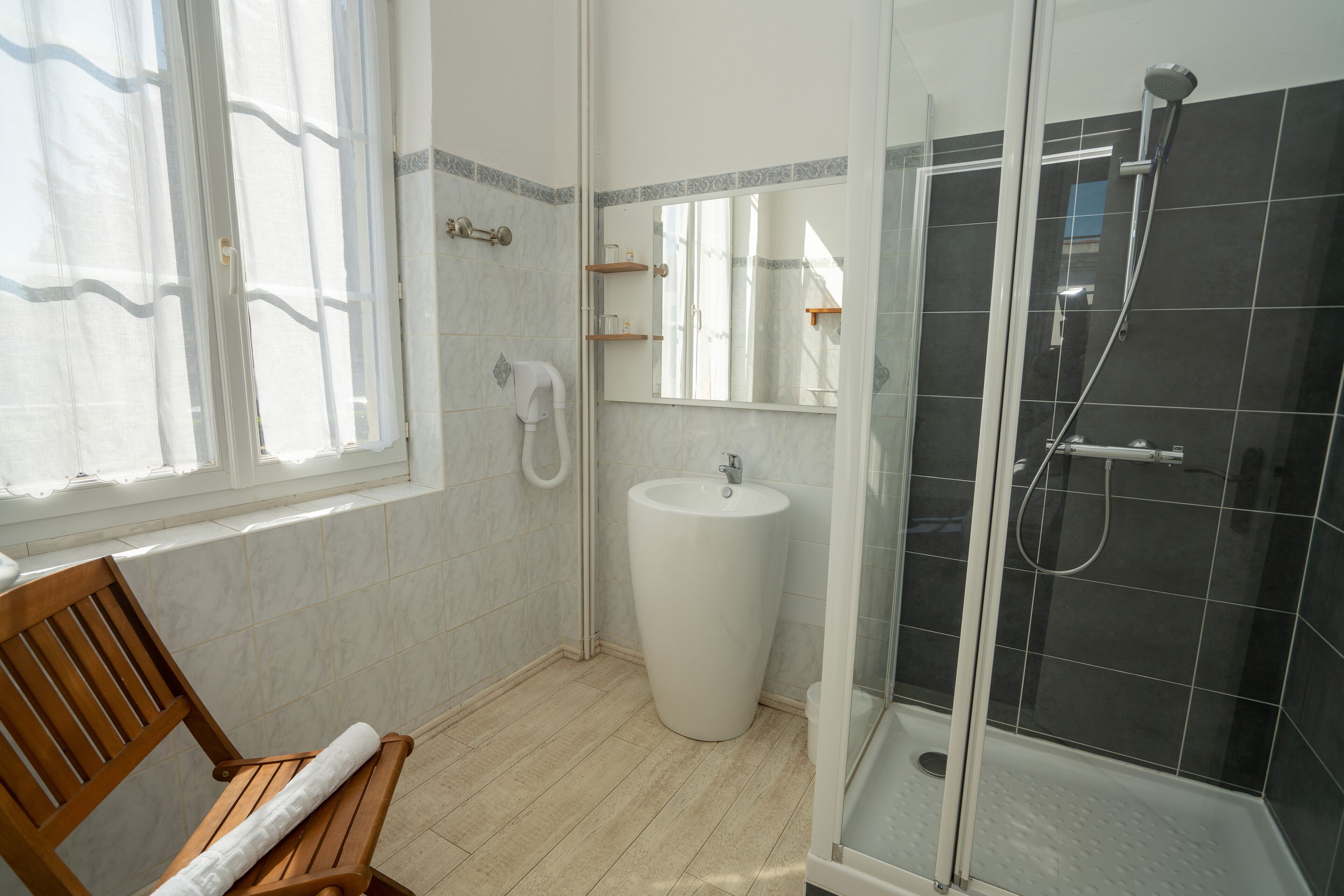 A bathroom with a wooden bench, a white sink, a glass shower enclosure, and a window with sheer curtains.