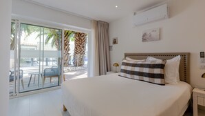 A hotel room with a bed, a nightstand, a wall-mounted air conditioner, a sliding glass door to a balcony, and a view of palm trees. - Hotel Le Square (Saint-Pierre-d'Oleron)