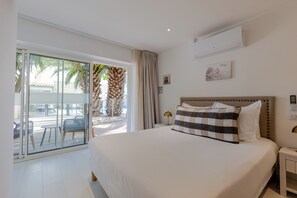 A hotel room with a bed, a nightstand, a wall-mounted air conditioner, a sliding glass door to a balcony, and a view of palm trees. - Hotel Le Square (Saint-Pierre-d'Oleron)