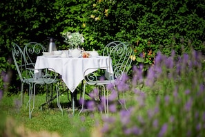 Une table dressée à l’extérieur, recouverte d’une nappe blanche, entourée de chaises en métal, et sur laquelle sont posés un pichet et une théière en métal.