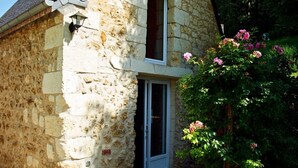 Een stenen huis met een schuin dak, een klein terras en een tuin vol bloemen en een boom.