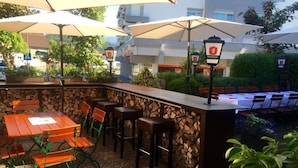 Un espace bar extérieur avec des tables et des chaises en bois, entouré de murs en pierre et abrité par des parasols.