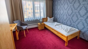 Une chambre individuelle avec un lit en bois, une chaise, une petite table, un radiateur et une fenêtre avec des rideaux.