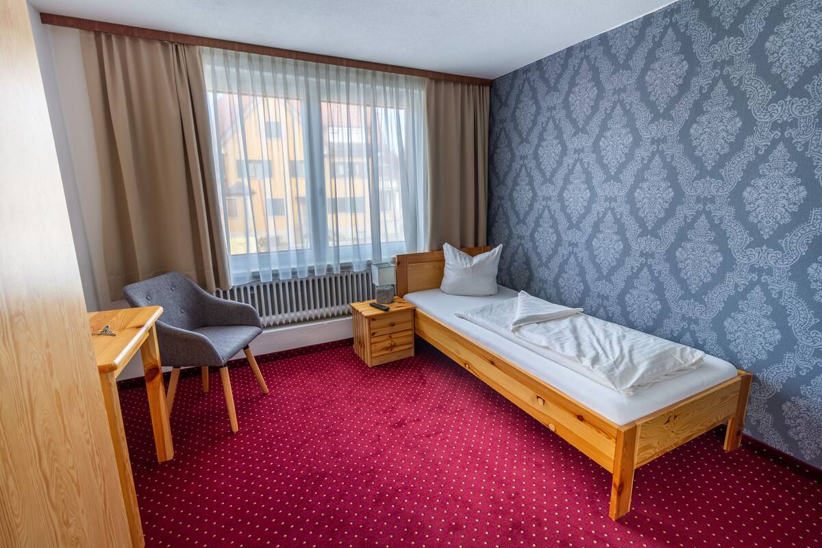 Ein Zimmer mit einem Einzelbett, einem Holzbett, einem Sessel, einem kleinen Tisch, einem Heizkörper und einem Fenster mit Vorhängen.