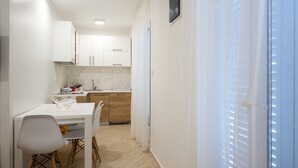 A compact kitchen with white cabinets, a dining table with chairs, and a window with blinds. - Apartmani Mia (Sveti Stefan)