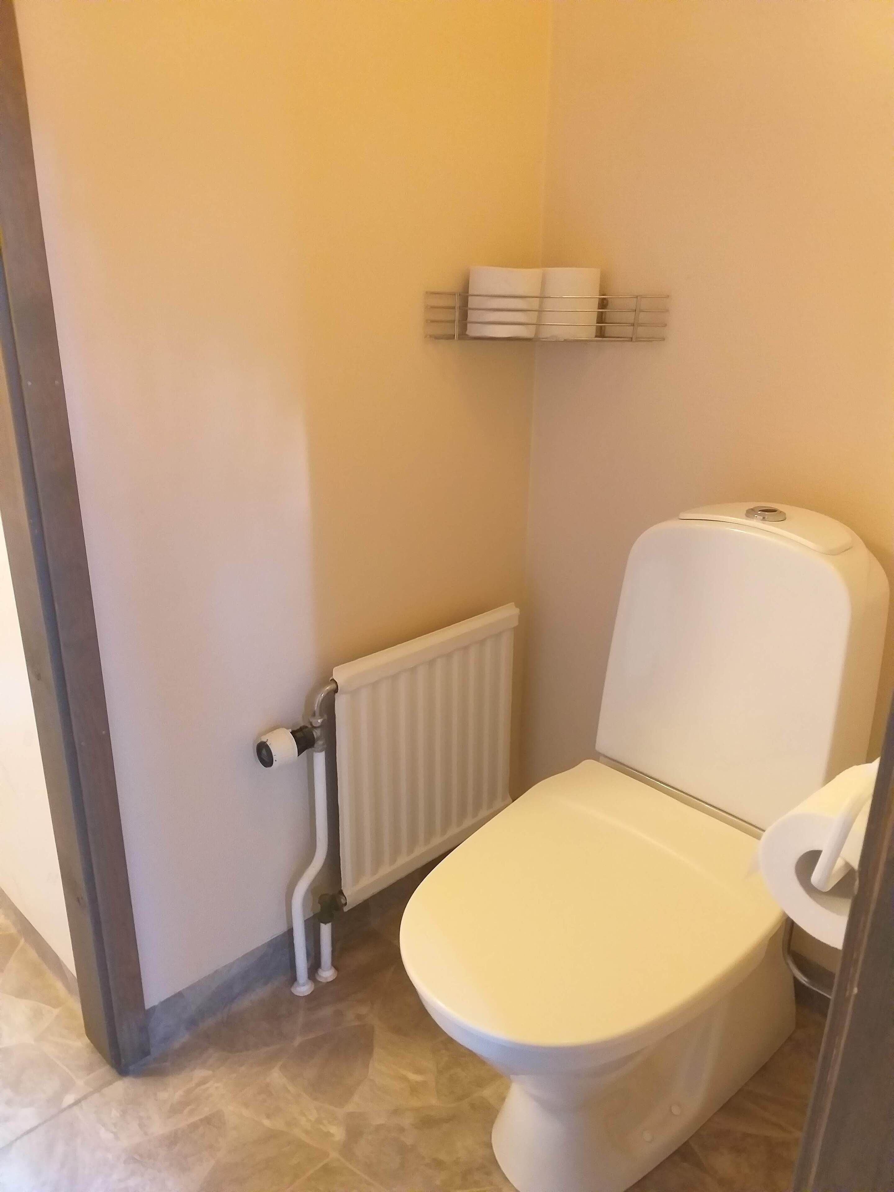 Une petite salle de bain propre, équipée de toilettes, d’un radiateur et d’une étagère avec du papier toilette.