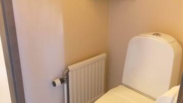 Une petite salle de bain propre, équipée de toilettes, d’un radiateur et d’une étagère avec du papier toilette.