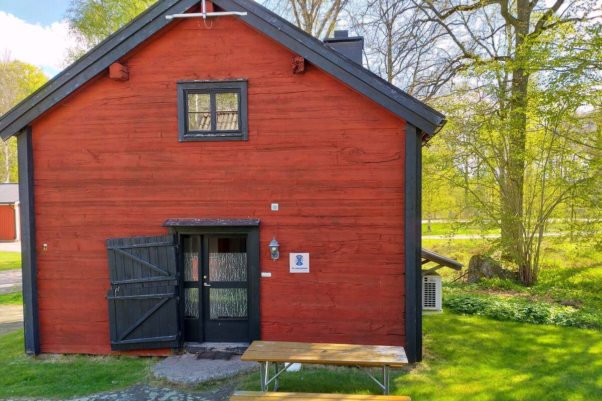 En rød træbygning med en sort dør og et vindue, et picnicbord foran og et skilt på væggen.