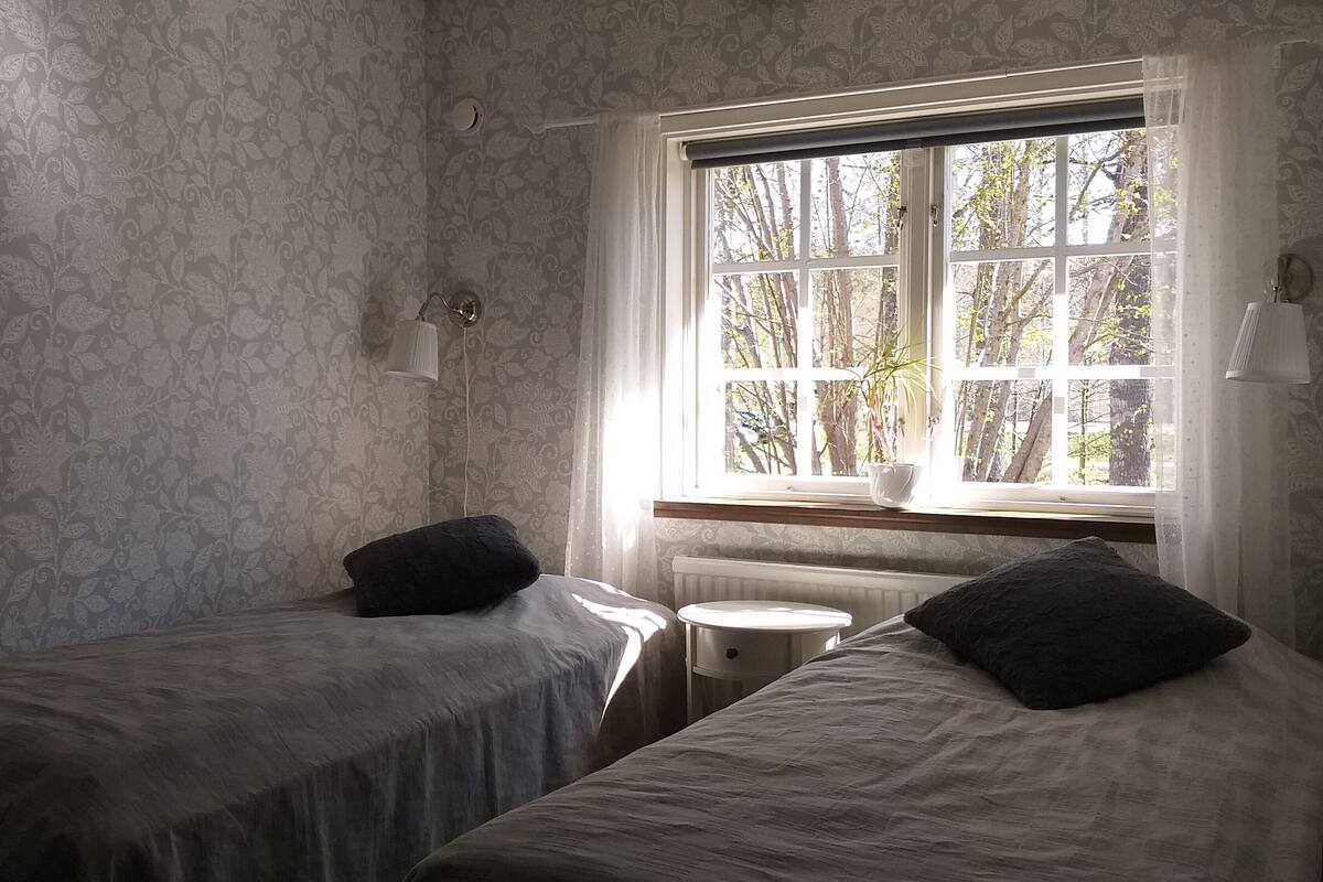 Et værelse med to enkeltsenge, en radiator, et vindue med udsigt til træer, og tapet med mønster.