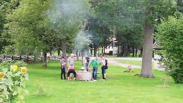 A group of people gathered around a barbecue grill in a park with trees and a wooden fence in the background.