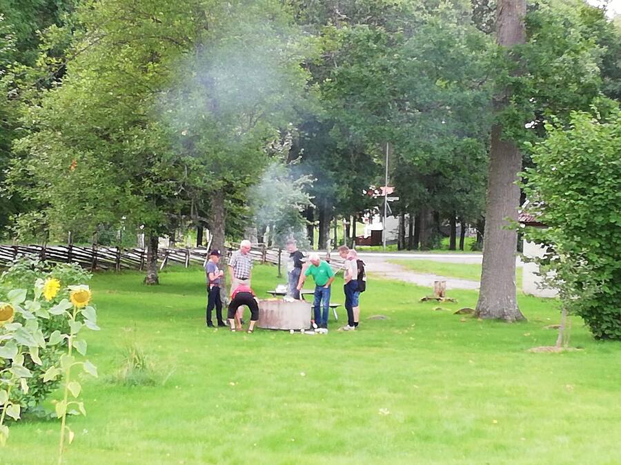 A group of people gathered around a barbecue grill in a park with trees and a wooden fence in the background.