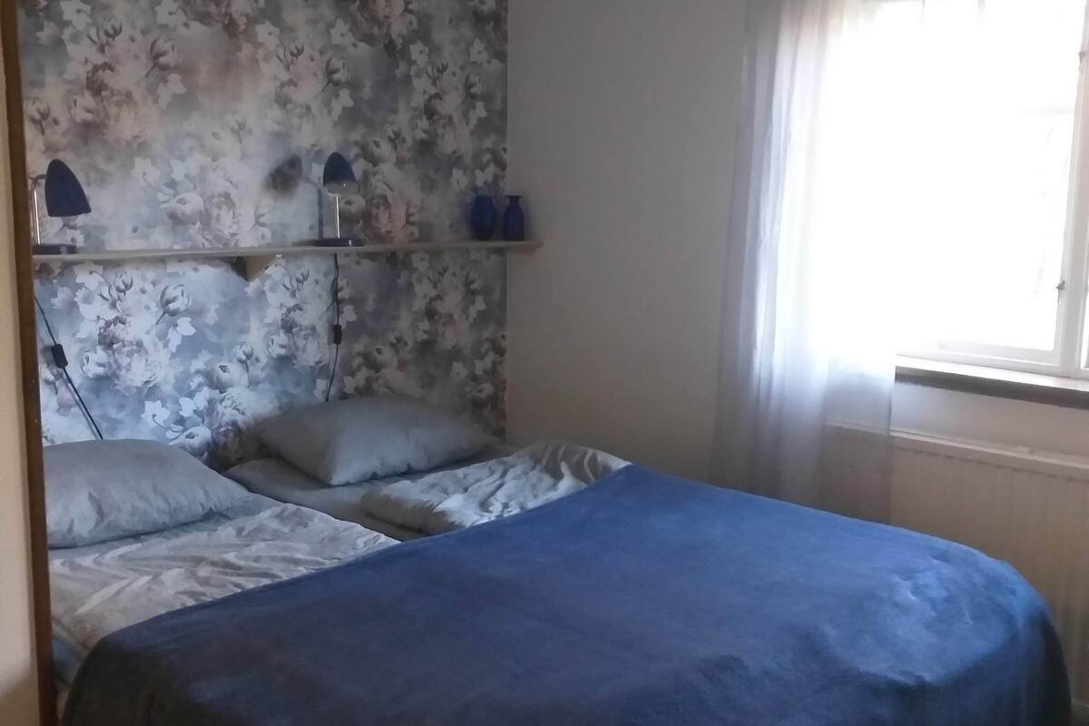 Et soveværelse med seng, blomstret tapet, et vindue med gardiner og en hylde med pynteting.