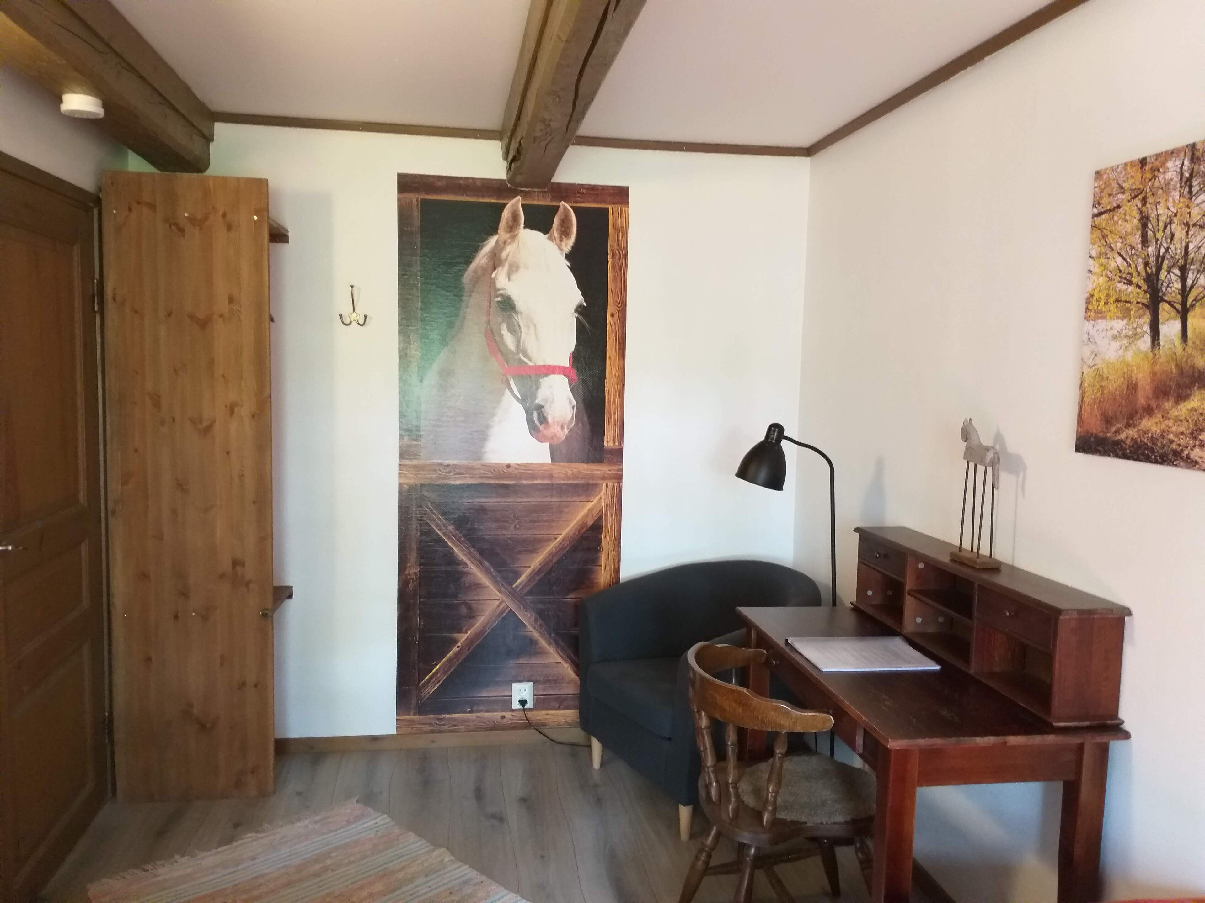 Una habitación con puerta de madera, un cuadro con tema de caballos, un escritorio con lámpara, una silla y una pequeña mesa.