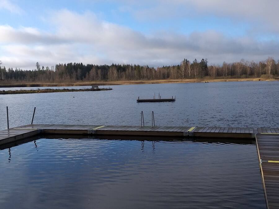 En träbrygga som sträcker sig ut i en stilla sjö, med en bänk och en liten båt i fjärran.