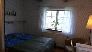 A small, cozy room with a bed, a chair, a table, and a window with a plant.
