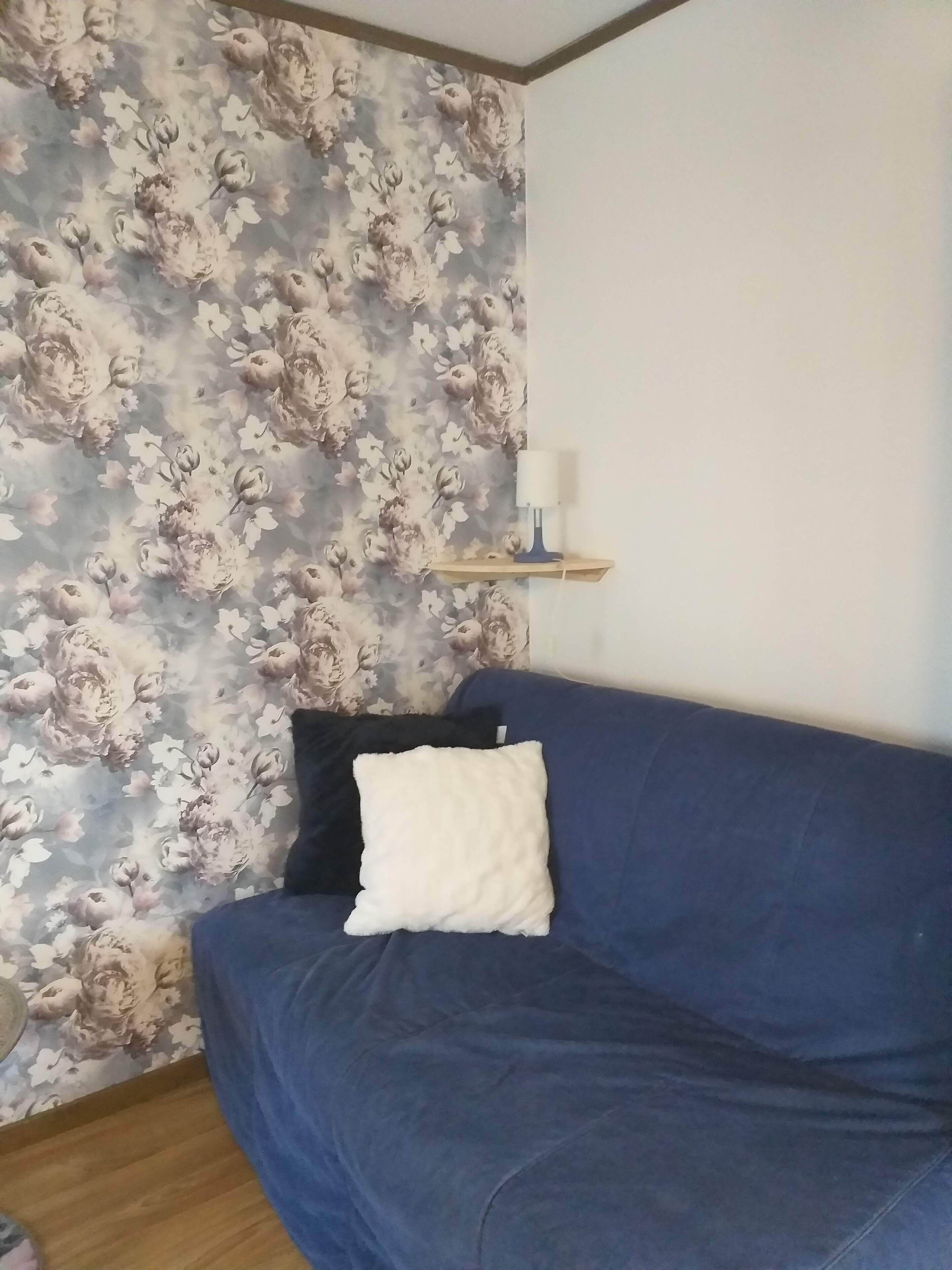 Une pièce avec un papier peint à motifs floraux, un canapé bleu avec un coussin blanc et une petite étagère sur laquelle se trouve une bougie blanche.