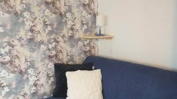 Une pièce avec un papier peint à motifs floraux, un canapé bleu avec un coussin blanc et une petite étagère sur laquelle se trouve une bougie blanche.