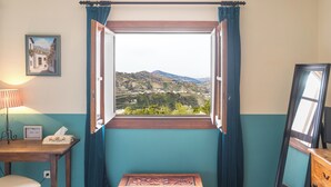 A room with a view of a mountainous landscape through an open window, a bedside table with a lamp, a framed picture on the wall, and a wooden chest.
