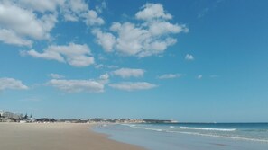 A wide sandy beach with gentle waves, a clear blue sky, and a lighthouse in the distance. - Residencial Sol (Portimão)