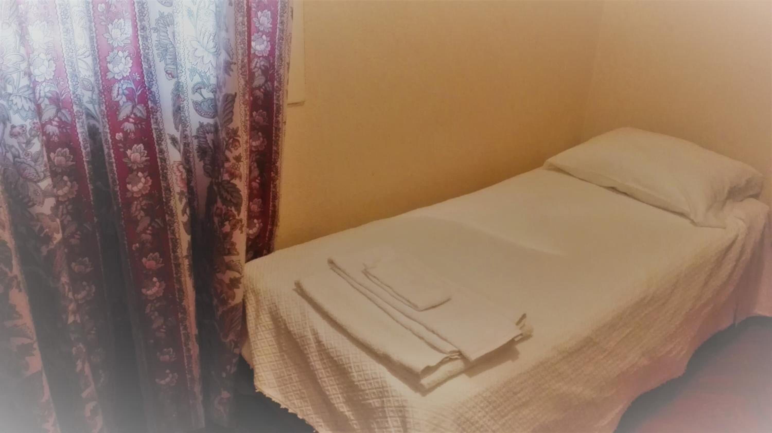 A small, single-bed room with a plain white bedspread and a single pillow. There is a patterned curtain partially drawn to the side.