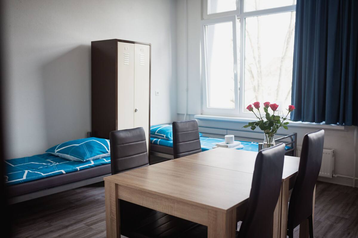 Ein Zimmer mit einem Bett, einem Tisch, Stühlen, einem Kleiderschrank und einem Fenster mit Vorhängen.