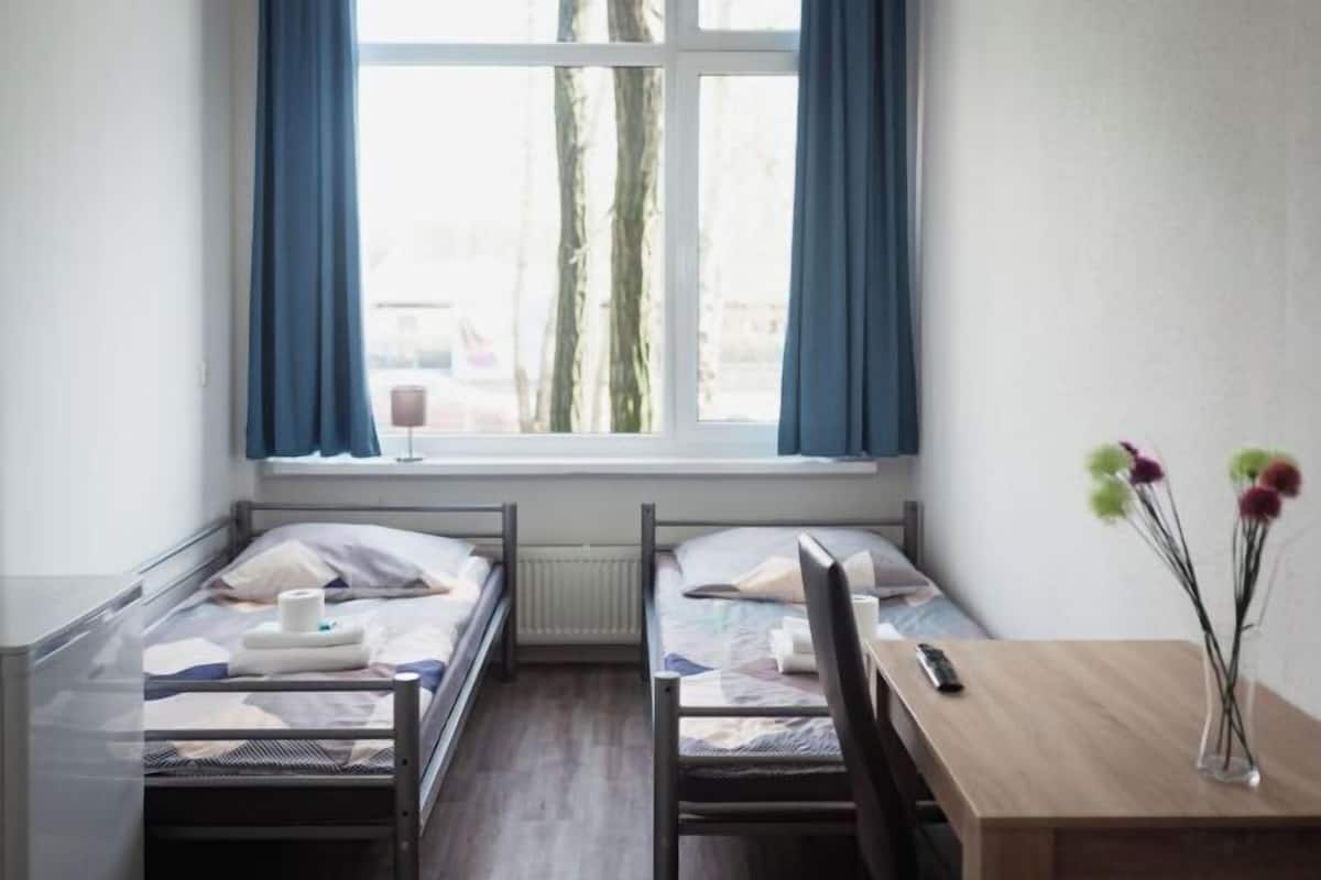 Ein Zimmer mit zwei Einzelbetten, einem Holztisch, einem Stuhl, einer Vase mit Blumen und einem Fenster mit Vorhängen.